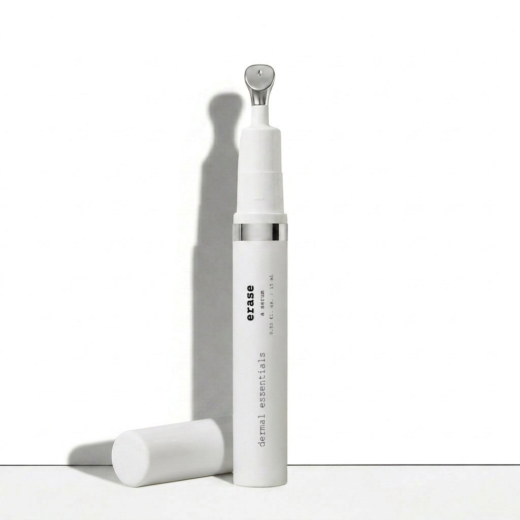 A white cylindrical skincare bottle labeled 'erase dermal essentials' with a silver metallic neck detail, shown standing upright with its cap placed beside it on a clean white surface.