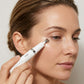 A high-resolution, close-up photograph capturing a woman of medium-light complexion with brown eyes and pulled-back hair, gently applying a white "Dermal Essentials - erase" eye serum pen with a silver applicator tip to the skin below her right eye. The white pen is held prominently in the foreground, showing legible black text. Her skin is moisturized and has a subtle, dewy sheen under warm, diffused light against a simple, light-colored backdrop.